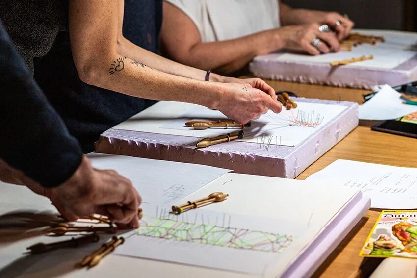 Tres pares de manos trabajan encaje de bolillos con hilos verdes, amarillos y naranjas en el museo